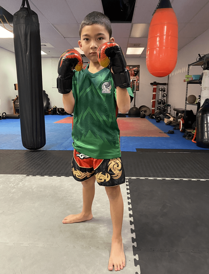 A young boy in green and black boxing gear posing in a martial arts gym with gloves and shorts.