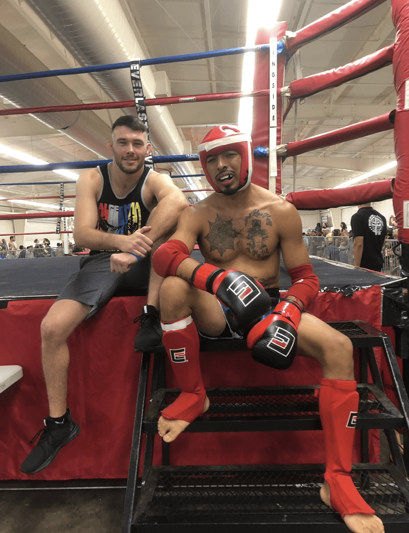 Two male boxers sitting in a boxing ring, one wearing red gloves and headgear, the other in casual sportswear.
