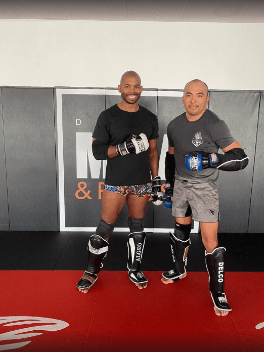 Two men wearing boxing gloves and protective gear standing in a gym, posing with fists raised.