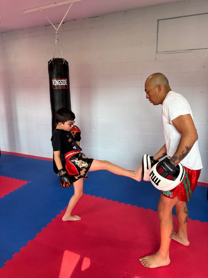 A young boy practices martial arts kicking a pad held by an adult trainer in a gym.