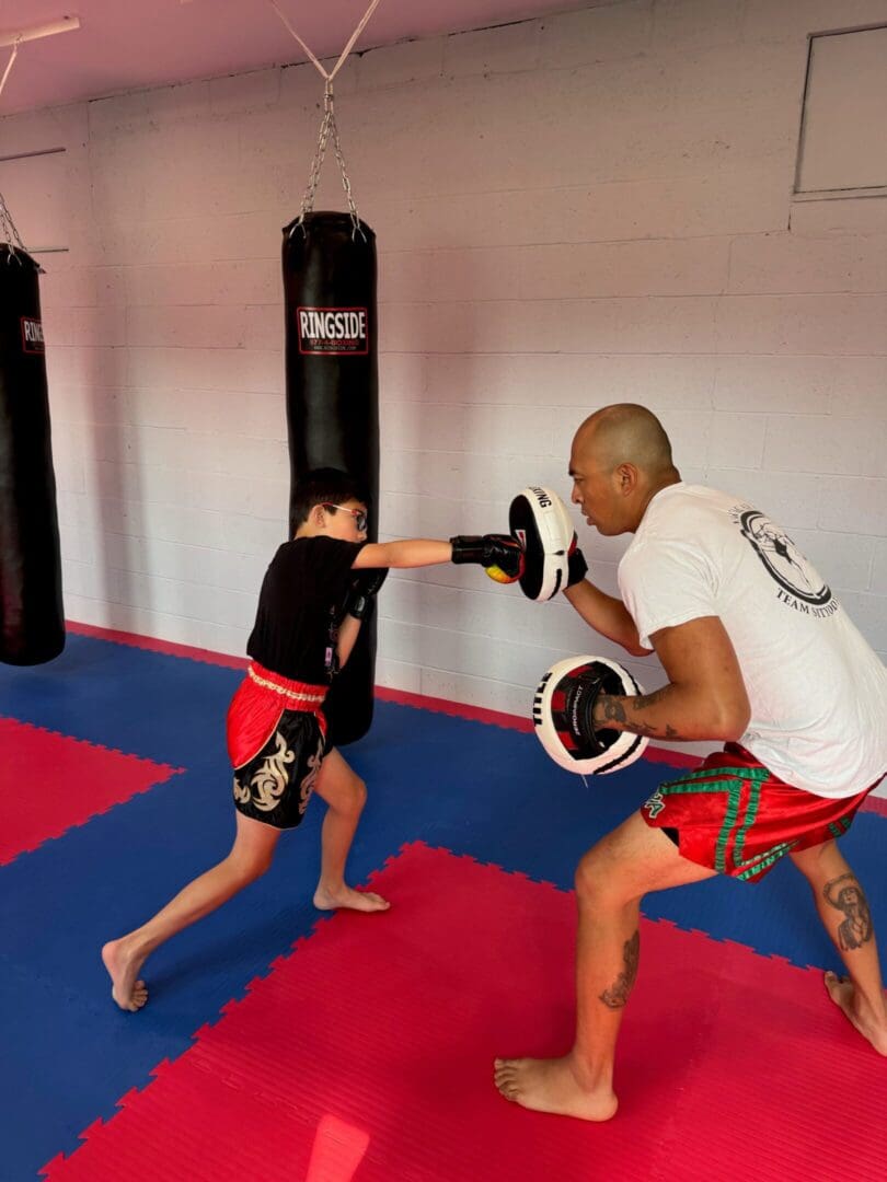 A young boy practices boxing punches with a trainer holding pads in a gym with red and blue mats.