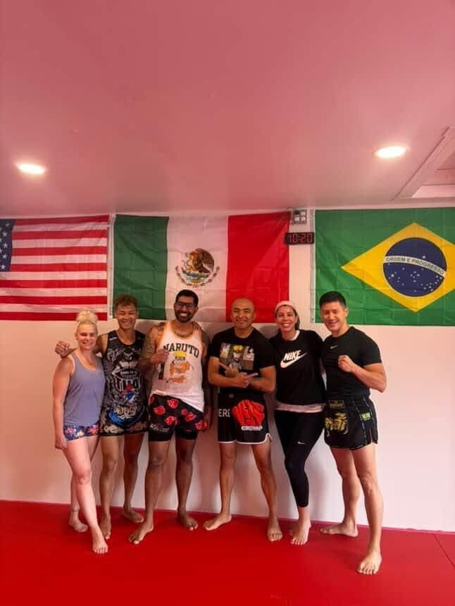 A group of six people posing in front of American, Mexican, and Brazilian flags in a gym setting.