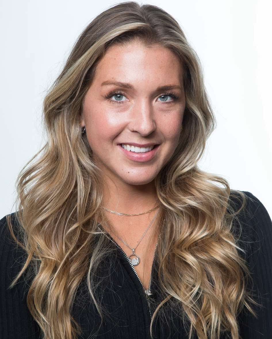 A smiling woman with long wavy blonde hair wearing a black top against a white background.